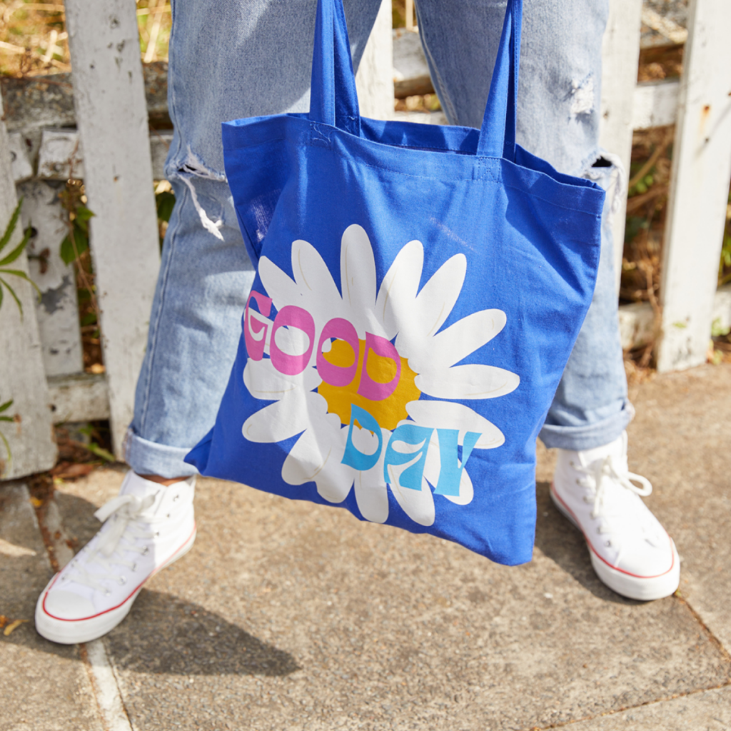A person in white sneakers and ripped jeans holds a blue tote bag with a white daisy and "Good Day" text, against a sunny, outdoor backdrop.