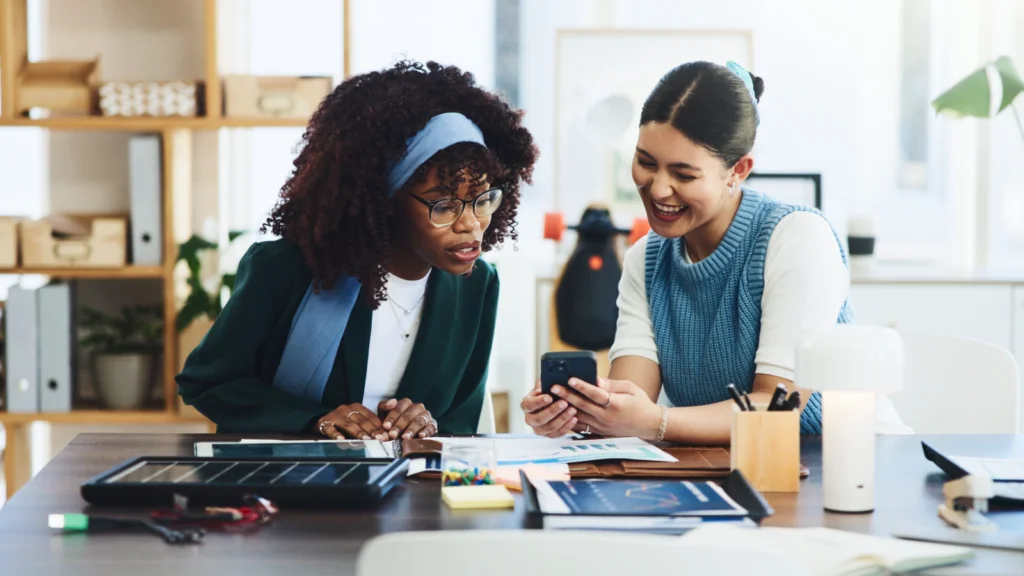 Develop simple AI tools or apps Two women in an office setting share a laugh while looking at a smartphone. The desk is scattered with papers and stationery.