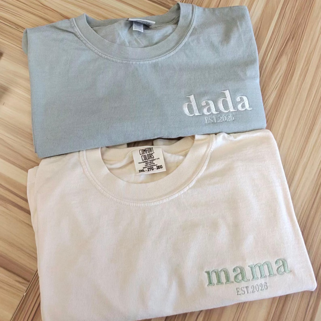 Two folded embroidered t-shirts on a wooden surface. The blue shirt reads "dada EST. 2026" and the white one reads "mama EST. 2026."