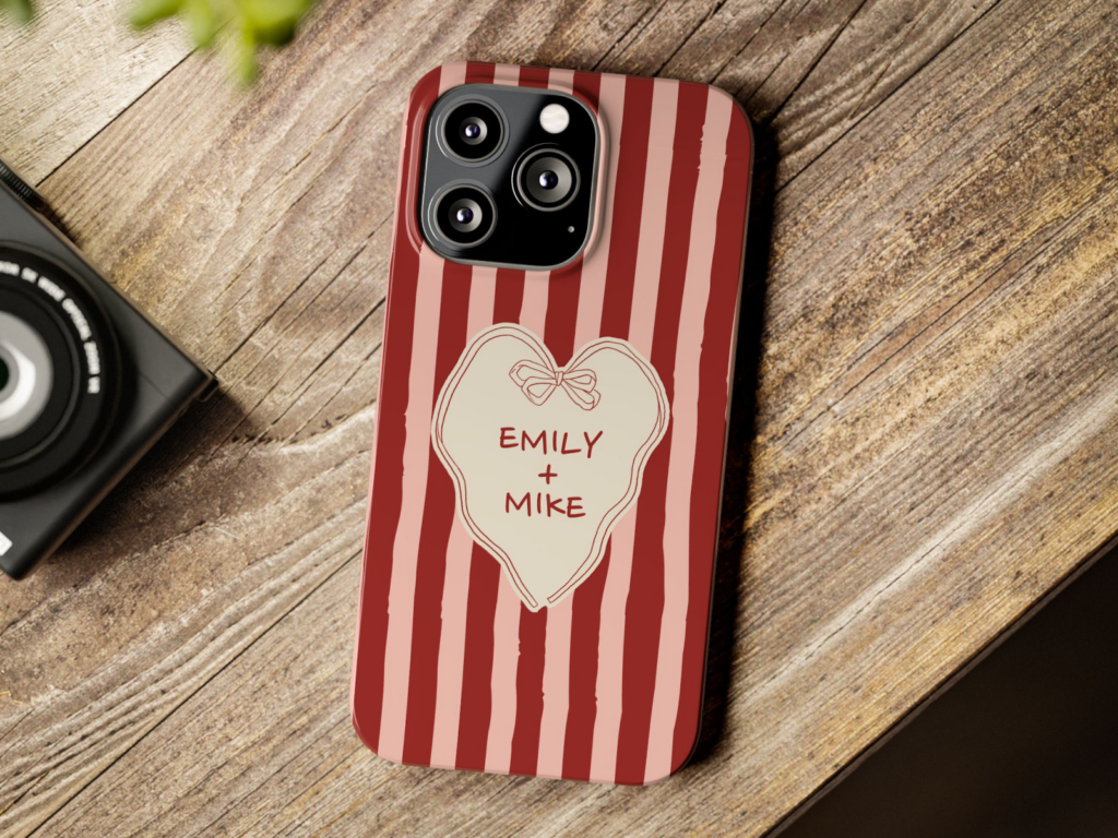 A phone with a red and pink striped case lies on a wooden surface. The case features a heart with "Emily + Mike" inside.