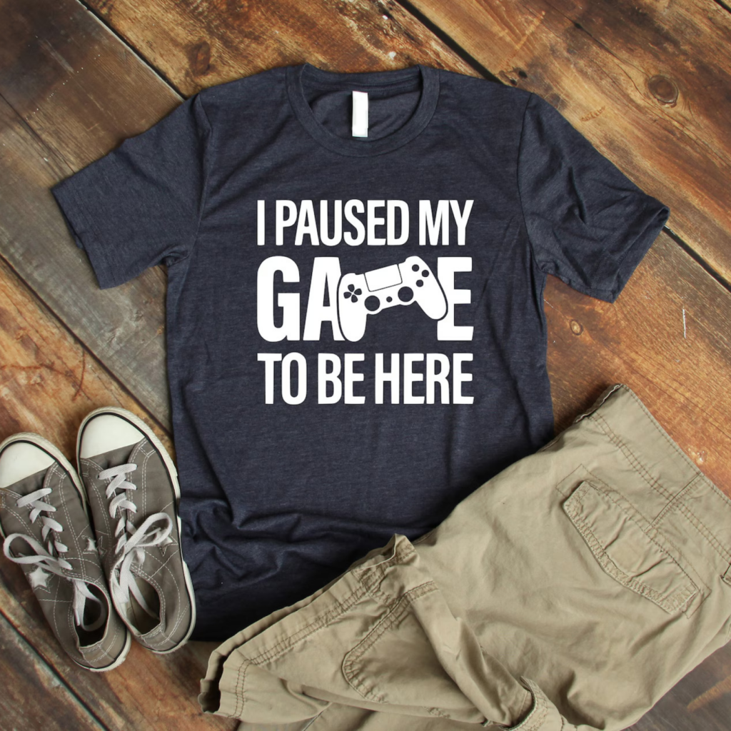 Navy t-shirt with "I Paused My Game to Be Here" and game controller graphic. Paired with beige shorts and gray sneakers on a rustic wood floor.