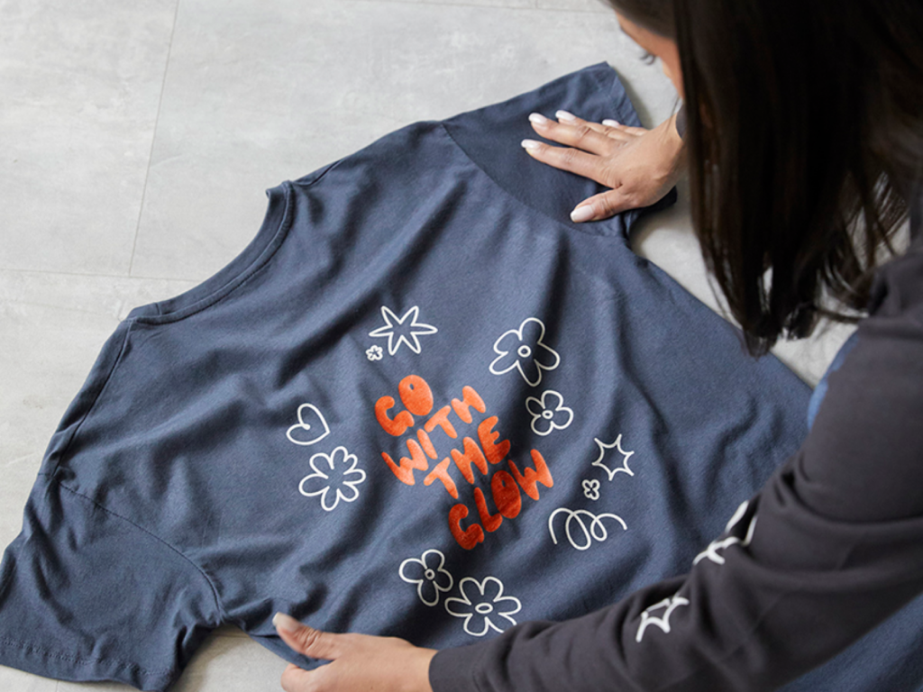 A woman arranges a blue t-shirt with "Go With The Glow" in bold orange text, surrounded by white flower and heart doodles.