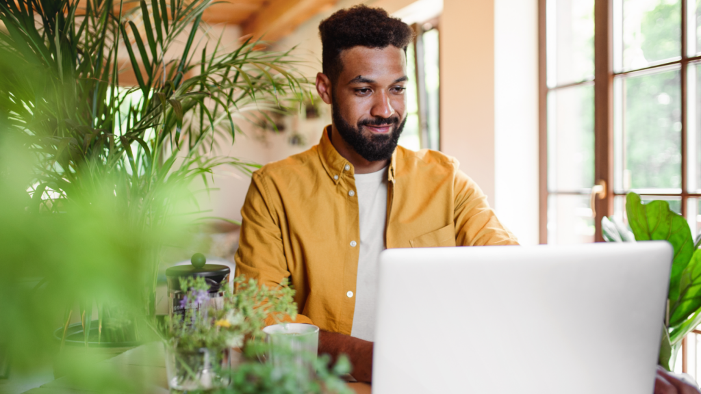 A man in a yellow shirt is working on a laptop, surrounded by lush green plants in a sunlit room.