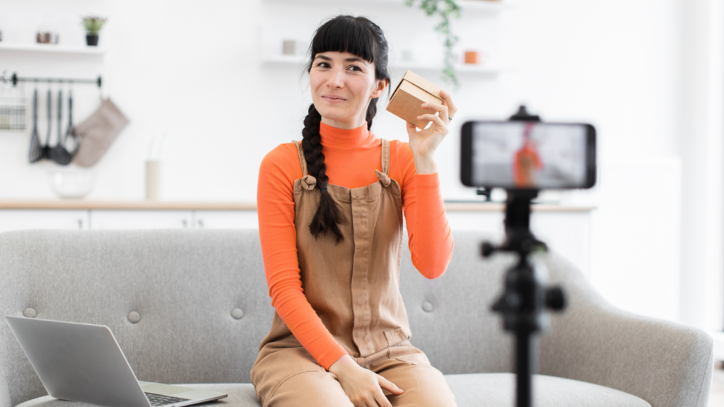 A woman with a braid smiles, holding a small box, sitting on a gray sofa. She's in front of a smartphone on a tripod, recording herself.