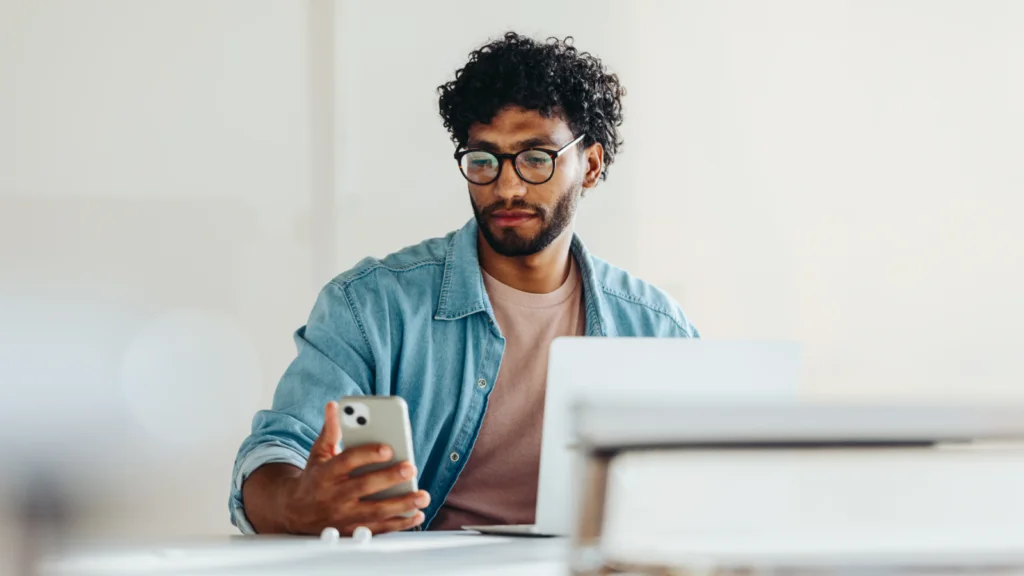 How to start making money with AI Step by step A man with glasses and curly hair looks at his smartphone while sitting at a desk with an open laptop.
