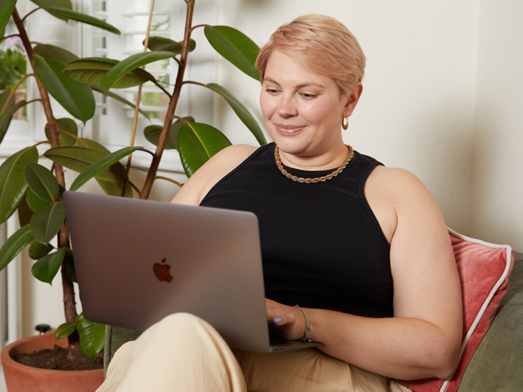A woman with short hair smiles while using a laptop on a sofa. She wears a black sleeveless top. A large potted plant is in the background.