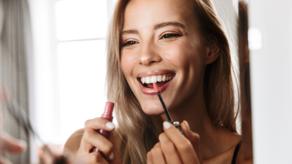 A smiling woman applies pink lip gloss while looking in a mirror.