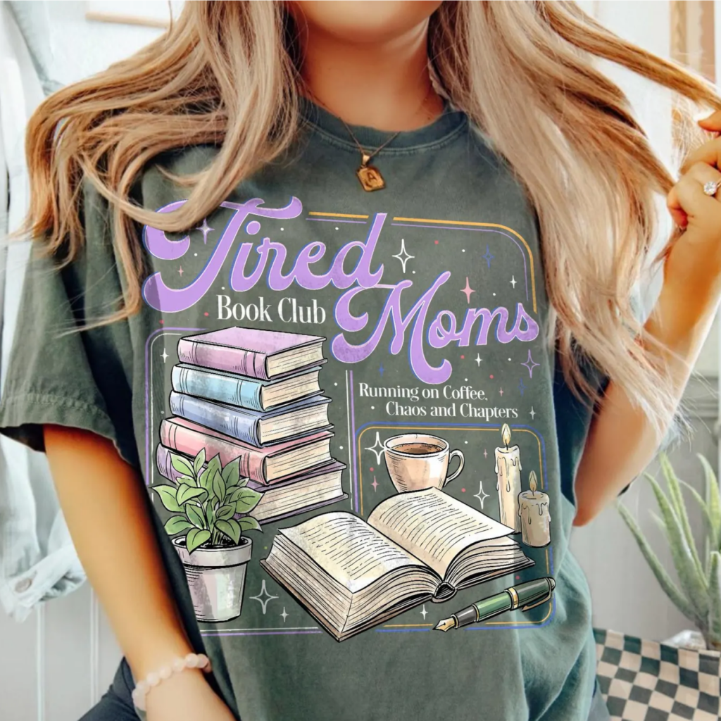 A woman in a dark green t-shirt with "Tired Moms Book Club" in purple letters. Illustrated books, plant, coffee cup, candles, and pen are displayed.