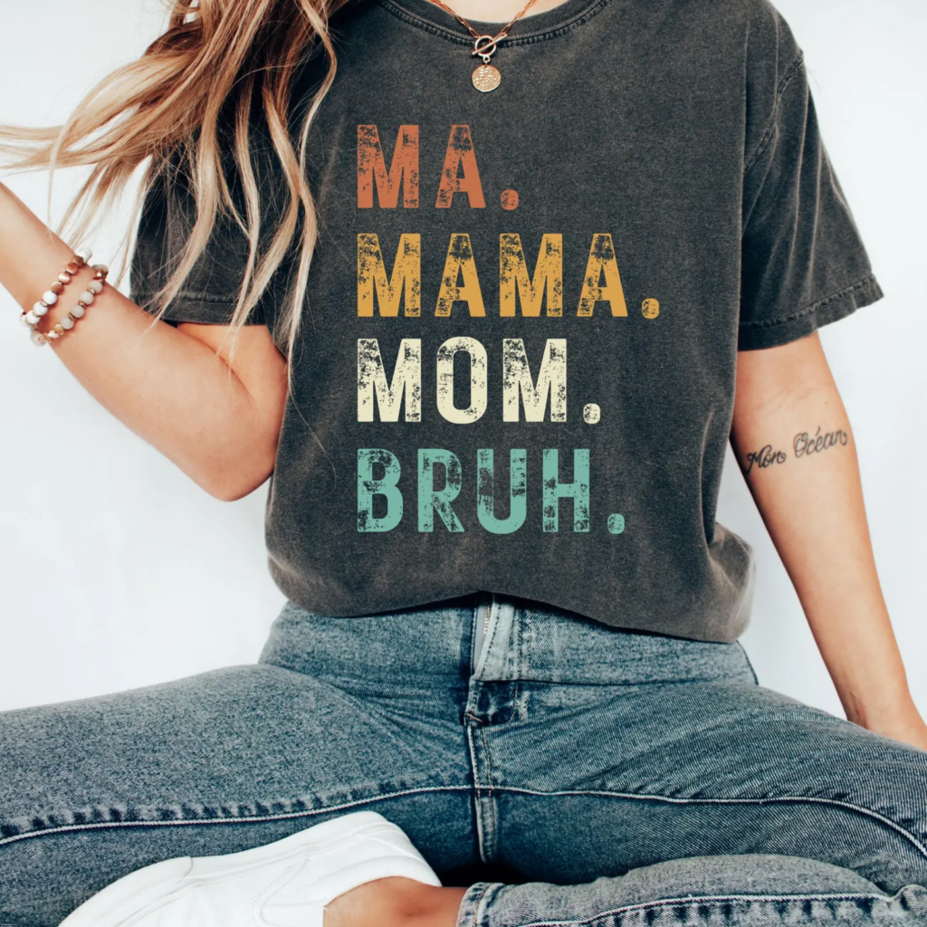 A woman wearing a black t-shirt with the words "MA. MAMA. MOM. BRUH." in colorful text. She's sitting cross-legged.