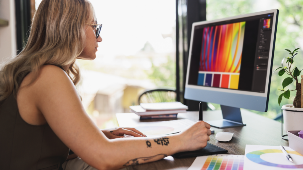 A woman with glasses uses a pen tablet at a desk, editing colorful digital art on a computer.