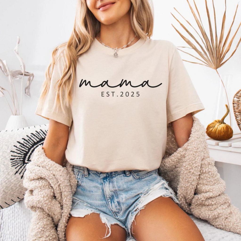 A woman wearing a beige "mama EST. 2025" t-shirt with denim shorts, sitting on a cozy chair.