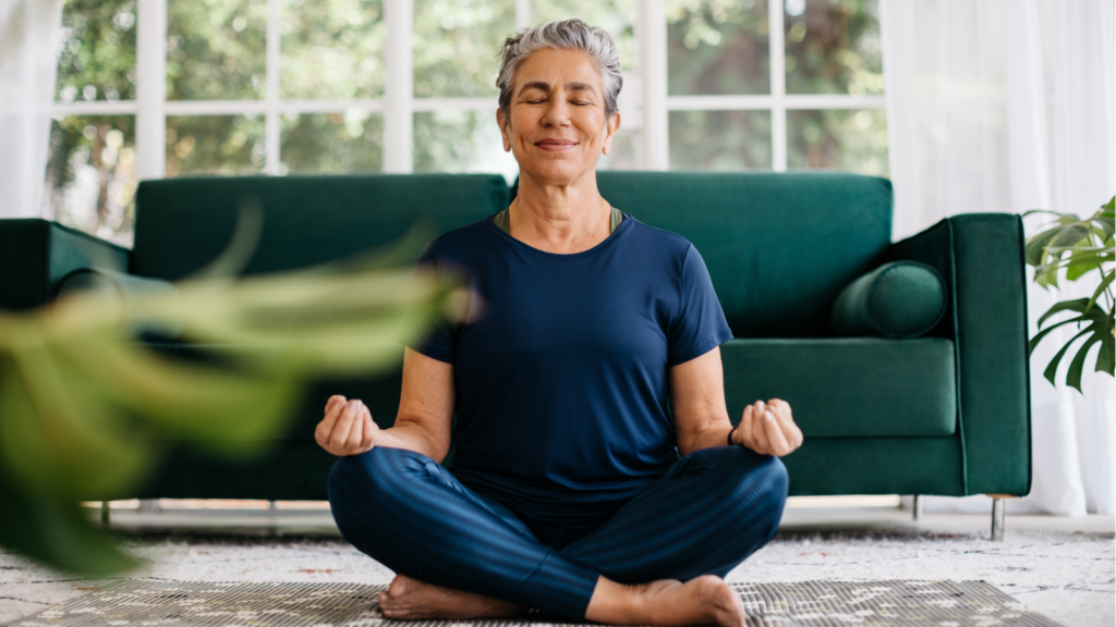 An older woman with short gray hair meditates in a serene, sunlit living room. She sits cross-legged on a mat, eyes closed, wearing a blue outfit.