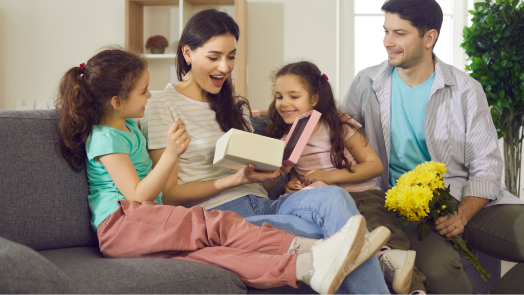 A joyful family scene with a mother and two daughters opening a gift on a sofa. The father, holding yellow flowers, smiles warmly.