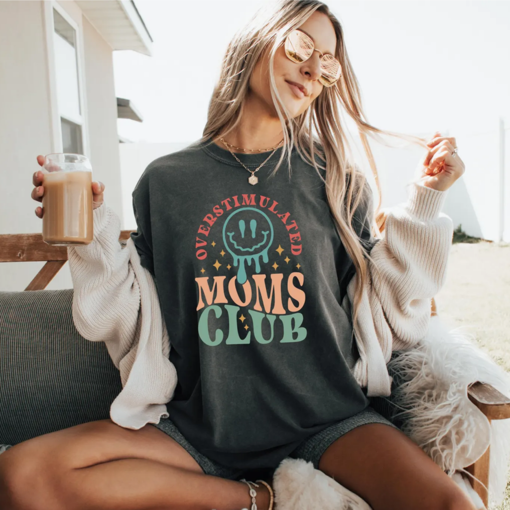A woman sits outdoors on a bench, wearing sunglasses and a "Overstimulated Moms Club" t-shirt. She holds an iced coffee.