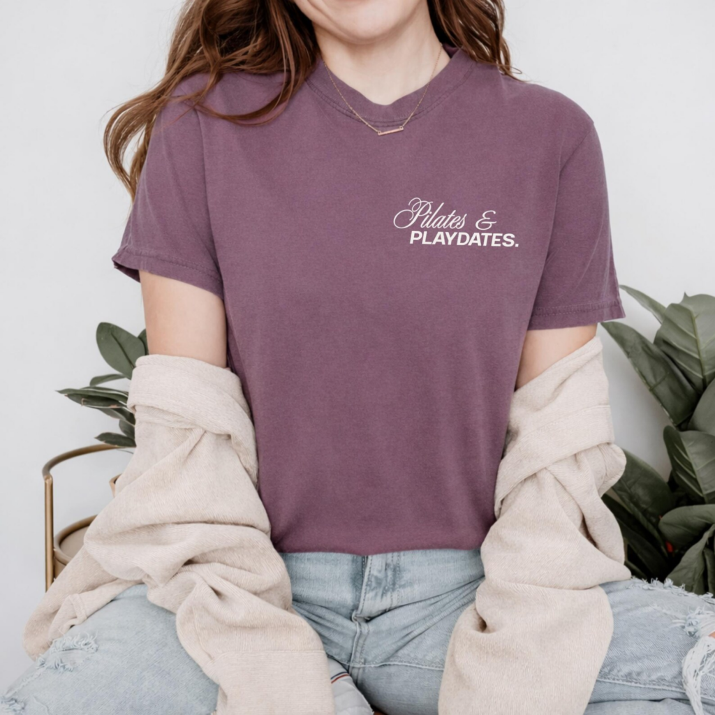 A woman wearing a mauve t-shirt with "Pilates & Playdates" text, light blue jeans, and a beige sweater draped over her shoulders, seated indoors.