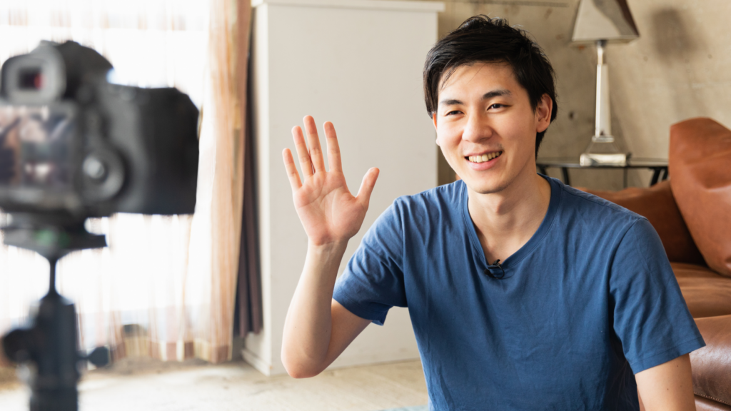 A smiling person in a blue shirt waves at a camera in a cozy room, as he records a video for YouTube.