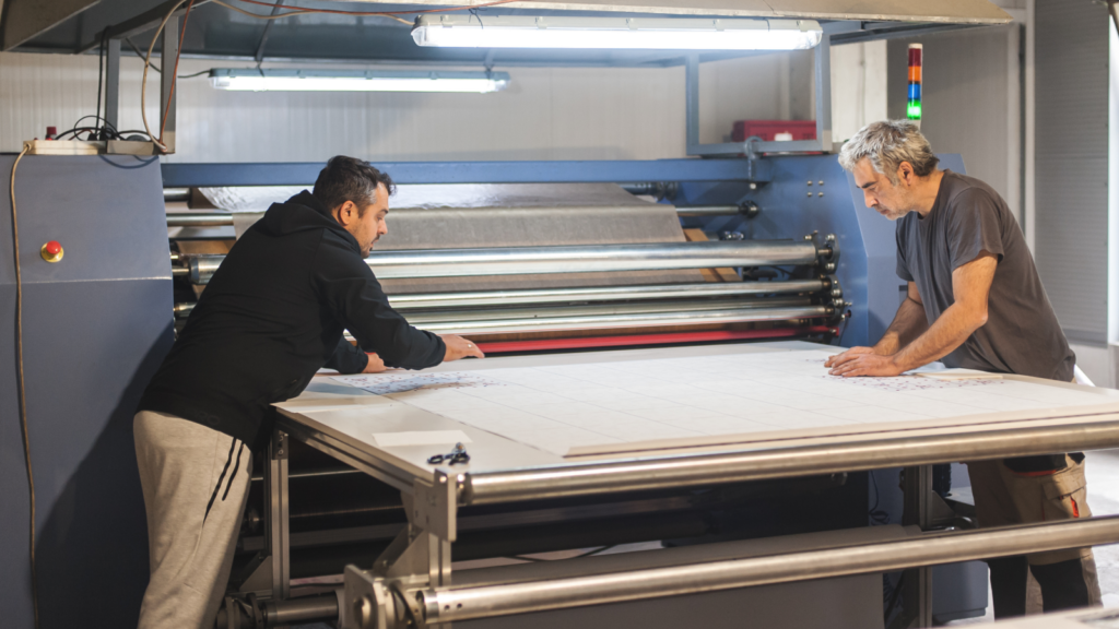 Two men focus intently on examining a large blueprint on a table in an industrial setting.
