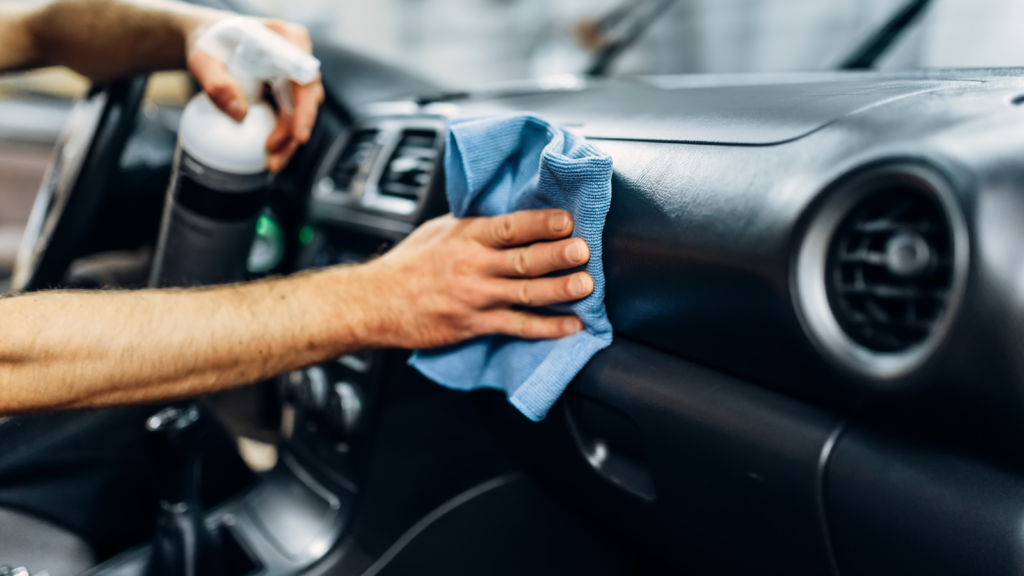 A person cleans a car dashboard with a blue cloth and a spray. The focus is on the hand and cleaning.