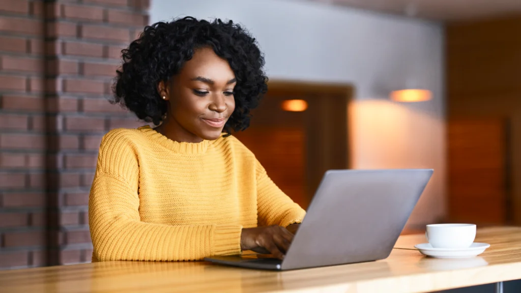 Sell digital products created with AI A woman in a yellow sweater uses a laptop to learn how to make money with AI at a wooden table with a cup of coffee nearby.