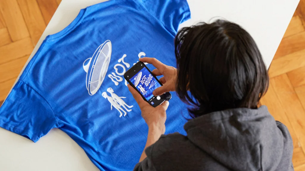 Sell print on demand products with AI A man in a hoodie photographs a blue t-shirt with a UFO design and the text "NOT ALONE" using a smartphone.