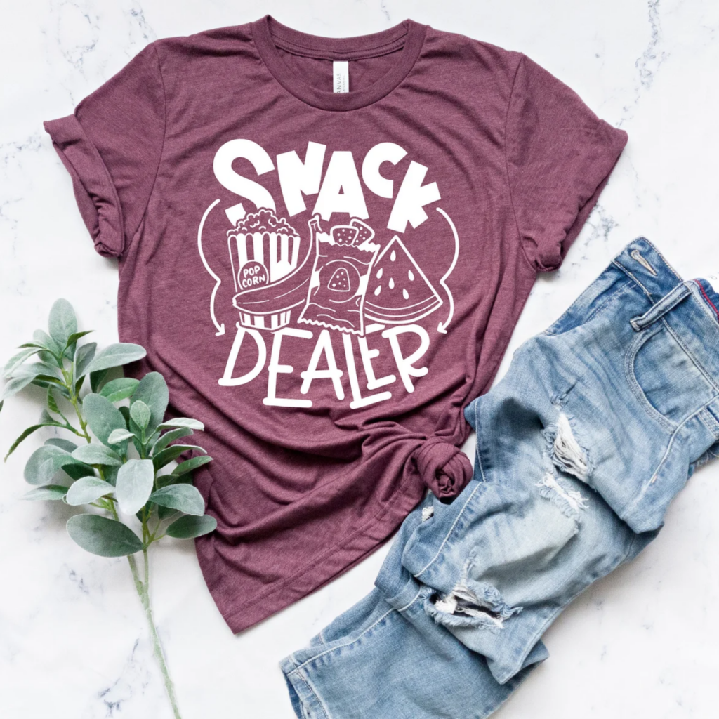 A maroon t-shirt with "Snack Dealer" printed in bold white letters, featuring popcorn, chips, and watermelon graphics.