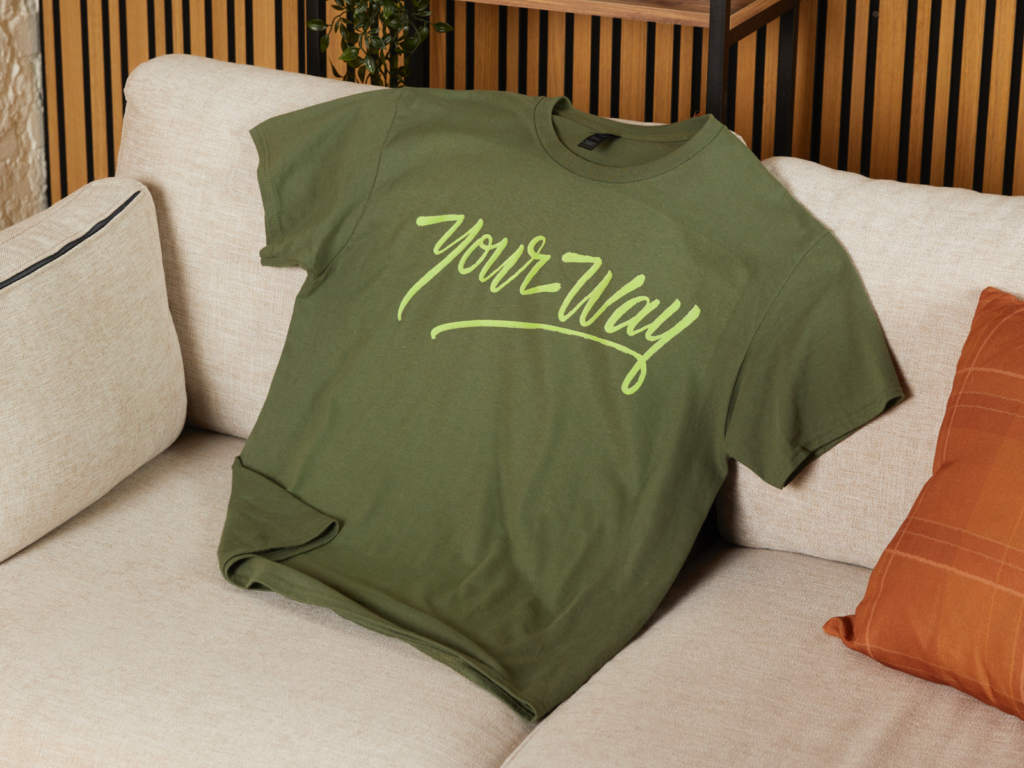 Green shirt with "Your Way" text in light-green laid on a light beige couch. Wooden backdrop and orange pillow behind it.