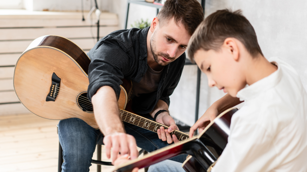 A man teaching a young boy to play guitar, both holding acoustic guitars. The setting is cozy and focused.