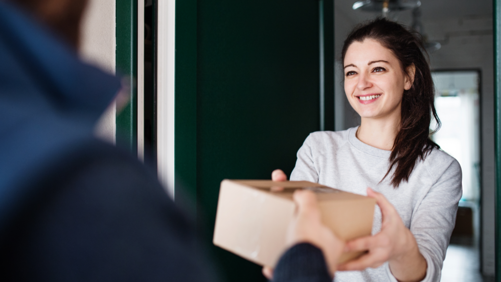 A smiling woman receives a package at her door from a delivery person. The setting is bright and welcoming, conveying warmth and positive interaction.