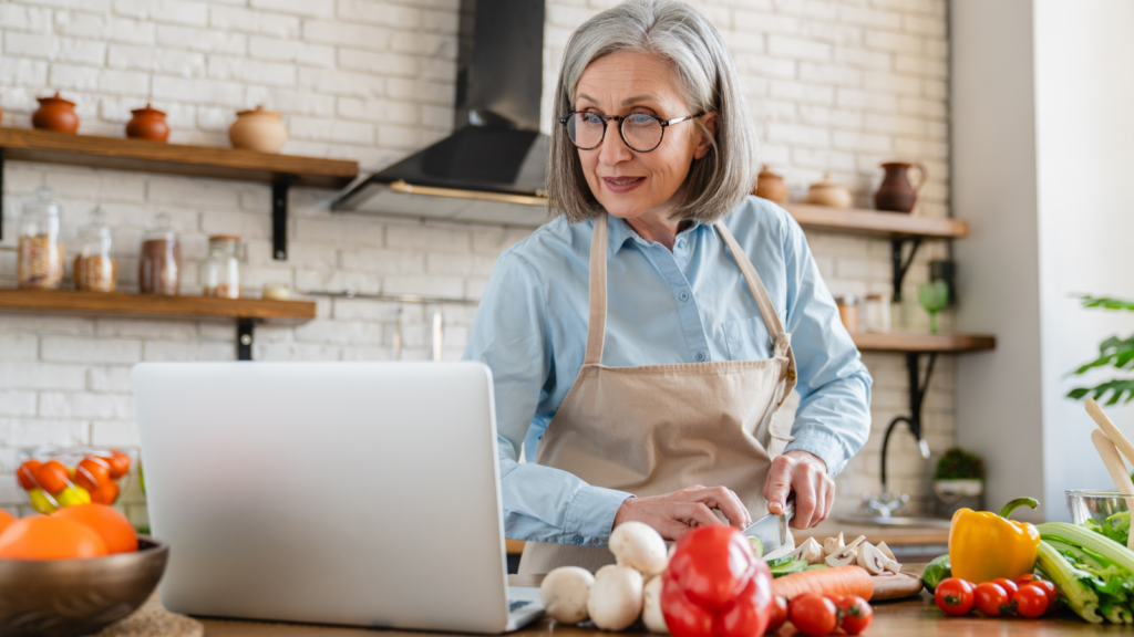 Elderly woman in an apron, smiling and chopping vegetables in a bright kitchen. She glances at a laptop, surrounded by fresh produce.