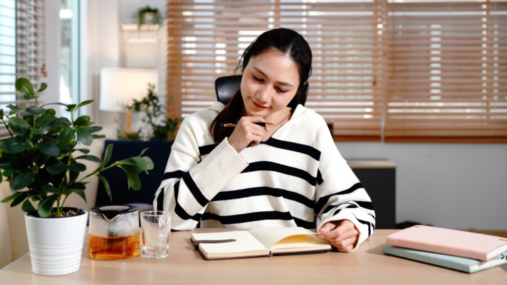 Woman in a striped sweater sits at a desk, thoughtfully writing in a notebook.