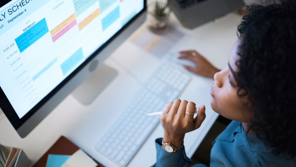 A woman sits at a desk, thoughtfully looking at a computer screen displaying a colorful weekly schedule.