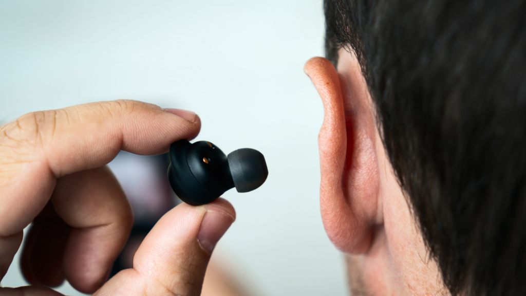 A man holding a black wireless earbud near their ear, preparing to insert it. The background is softly blurred.