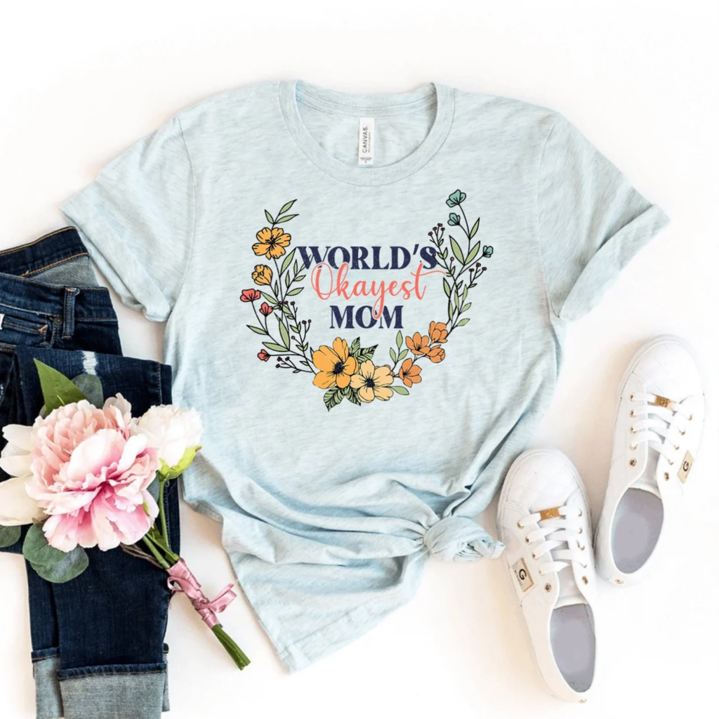 Light blue t-shirt with "World's Okayest Mom" in decorative font with colorful flowers. Paired with jeans, white sneakers, and a pink peony.