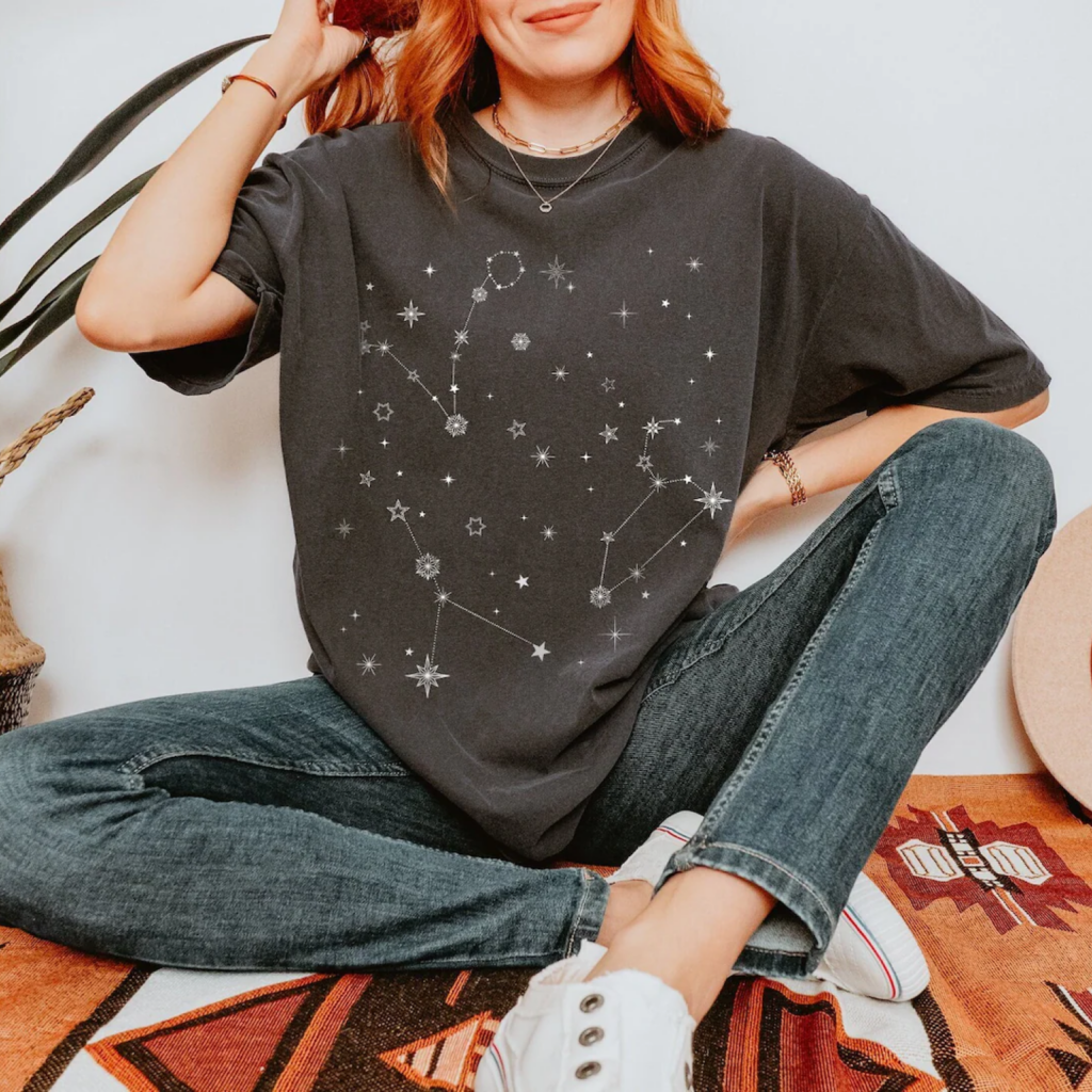 A woman in a dark t-shirt with star constellations print, sitting cross-legged on a colorful, patterned rug. Plants in the background.
