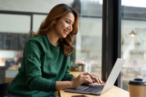 Smiling woman in a green sweater uses a laptop by a window in a cozy cafe.