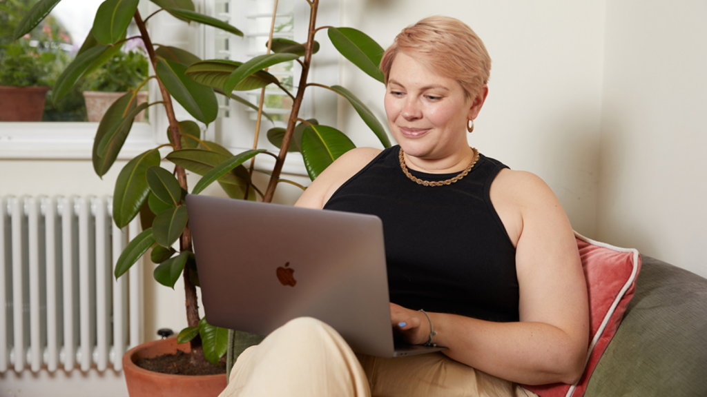 A woman with short hair sits on a couch, smiling at a laptop. She wears a black tank top and beige pants. Green plants and a window in the background.