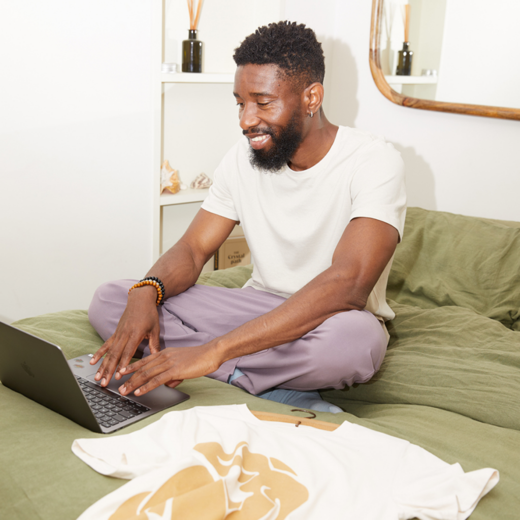 A smiling man is sitting on a bed with a laptop and a white t-shirt with a golden print in front of him.