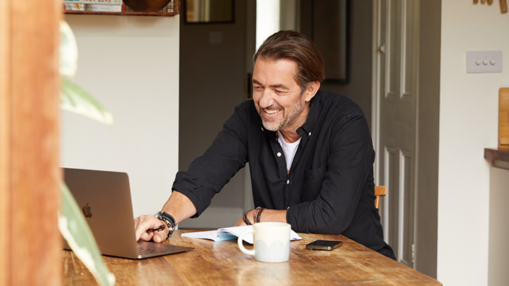 A smiling man in a blue shirt is using ChatGPT to create custom products with Printify (from a laptop). He’s sitting in a kitchen.