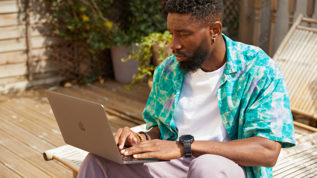 A man in a colorful shirt works on a laptop outdoors on a sunny wooden deck.