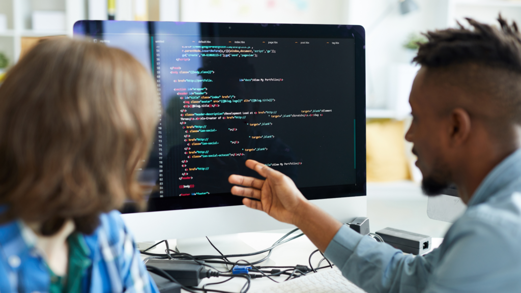 Two people are discussing code displayed on a computer screen. The person on the right gestures at the colorful lines of code.
