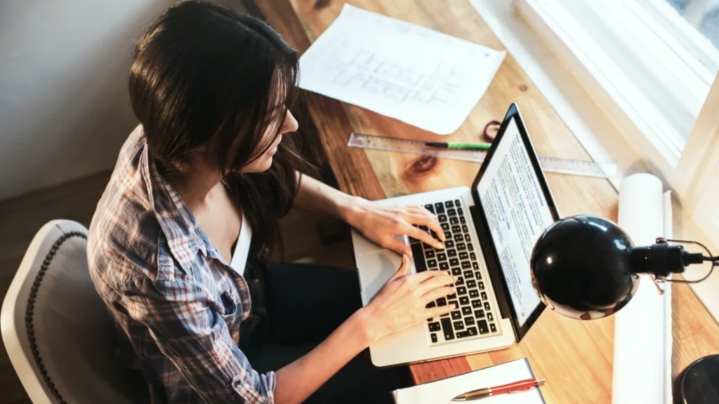 A woman in a plaid shirt works intently on a laptop at a wooden desk. Papers and a ruler are scattered, and a lamp illuminates the space.