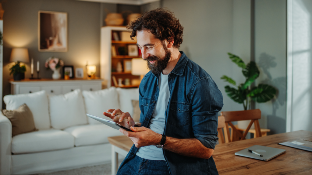 A man with curly hair and a beard sits in a cozy living room, smiling at a tablet. A sofa, plants, and decorative shelves in the background.