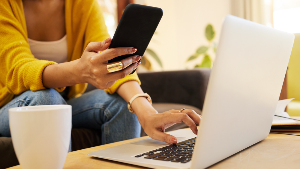 A person in a yellow cardigan uses a smartphone and a laptop at a wooden table.