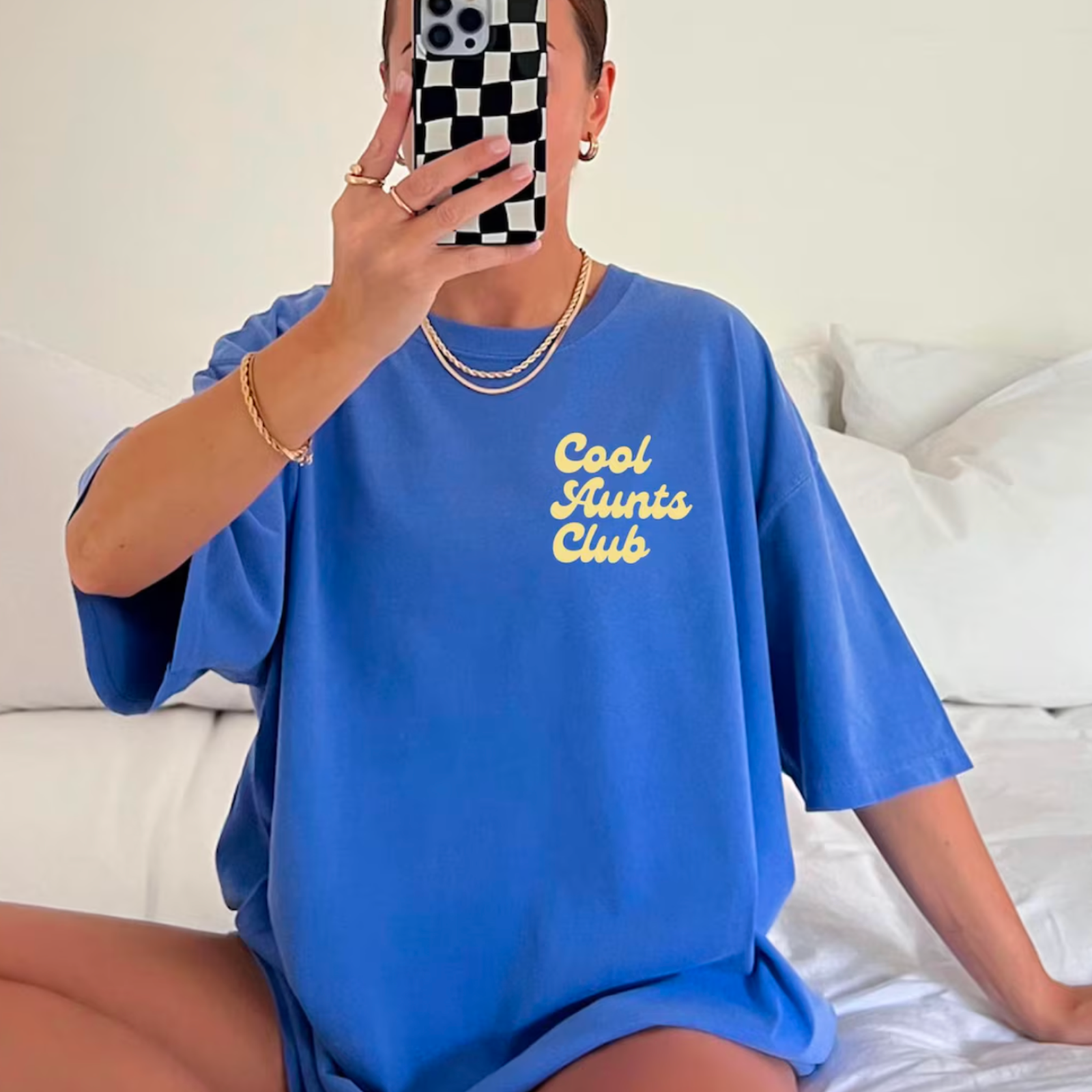 A person in a blue oversized shirt with "Cool Aunts Club" in yellow text takes a selfie on a bed.