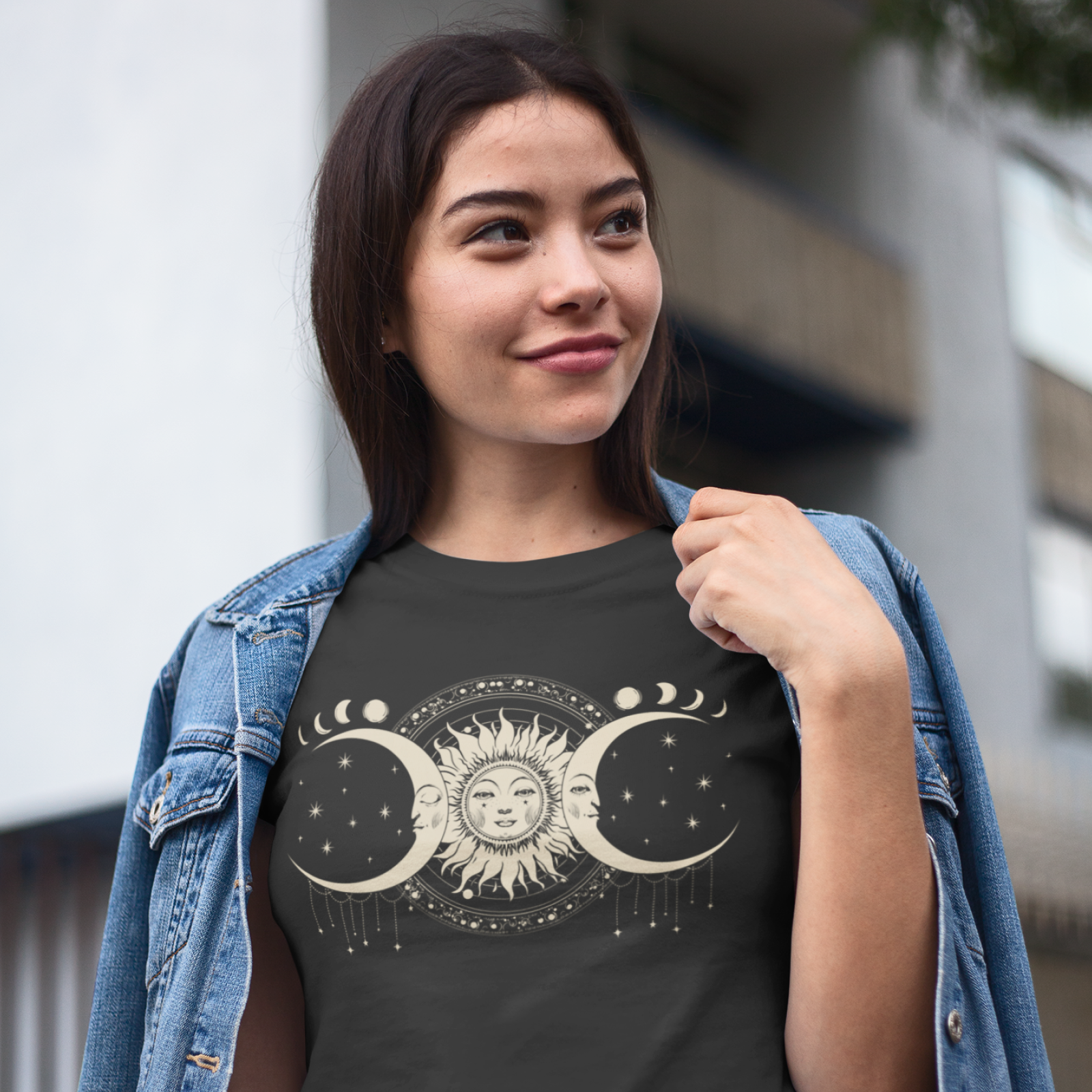 Young woman outdoors, wearing a denim jacket over a black T-shirt with a celestial design of the sun flanked by crescent moons, exuding a serene mood.