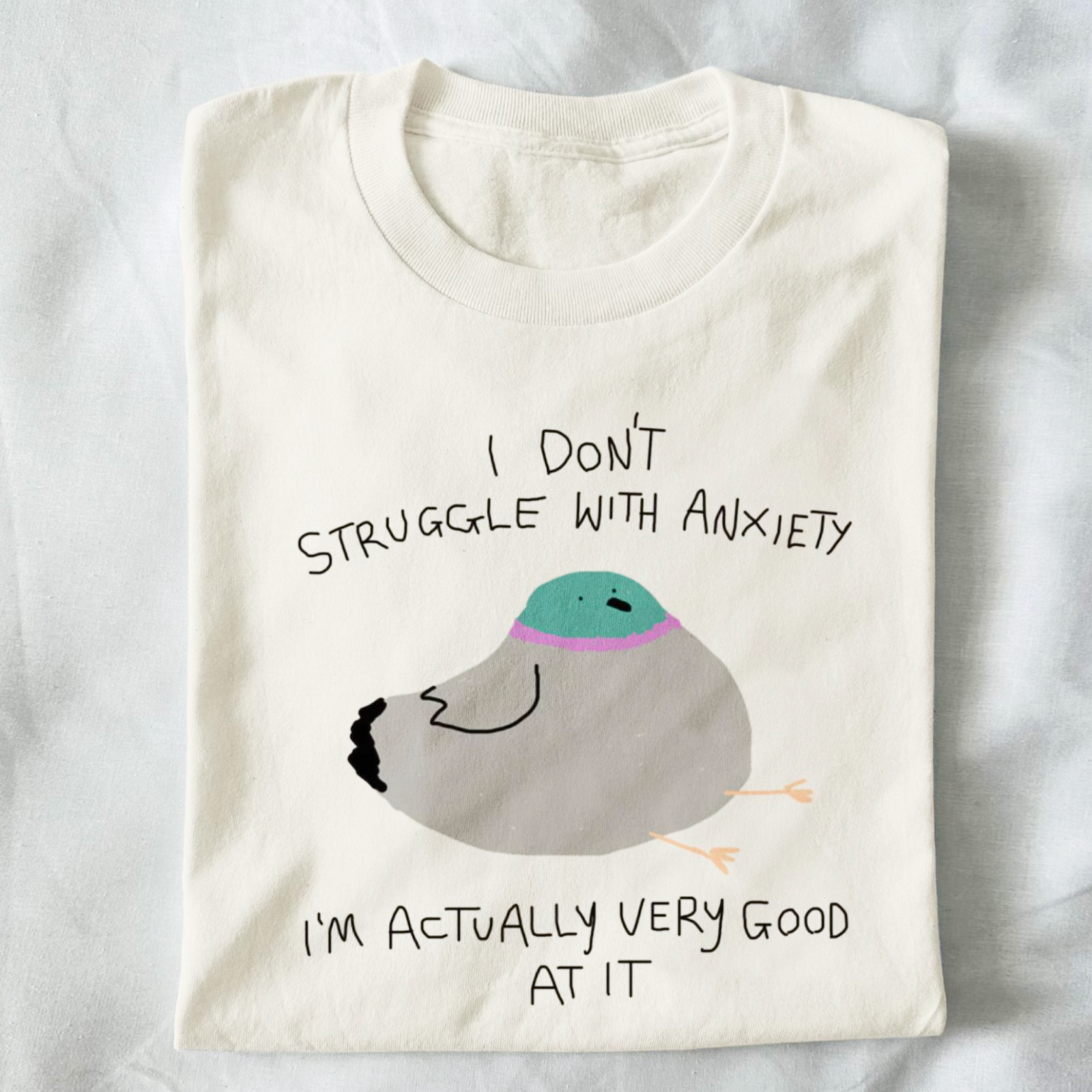 Folded cream t-shirt featuring a cartoon bird with teal head and purple band, captioned "I don't struggle with anxiety, I'm actually very good at it."