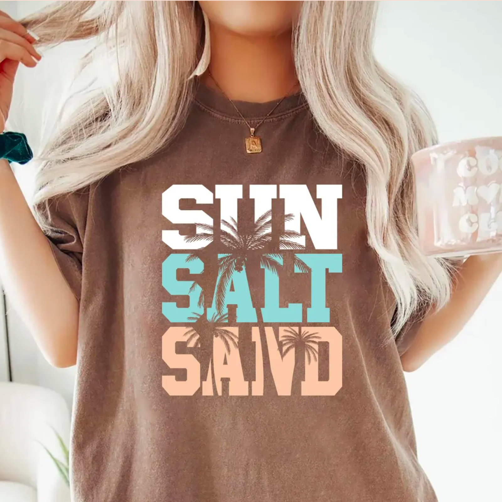 A woman with long blonde hair holds a mug and wears a brown shirt with the words "Sun Salt Sand" in bold white, turquoise, and peach letters.