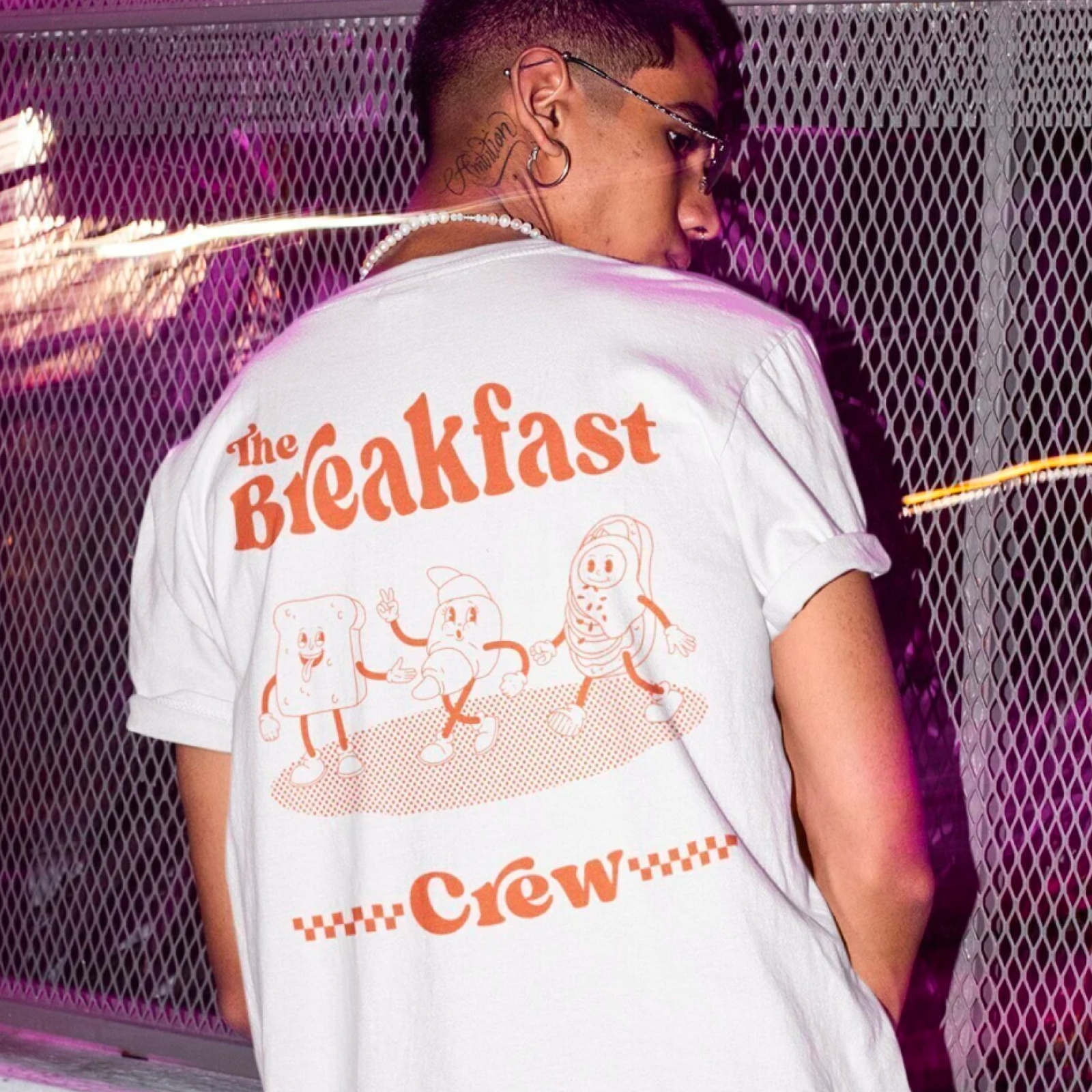A person in sunglasses faces away, wearing a white T-shirt with "The Breakfast Crew" and cartoon breakfast characters in orange. Urban, playful vibe.