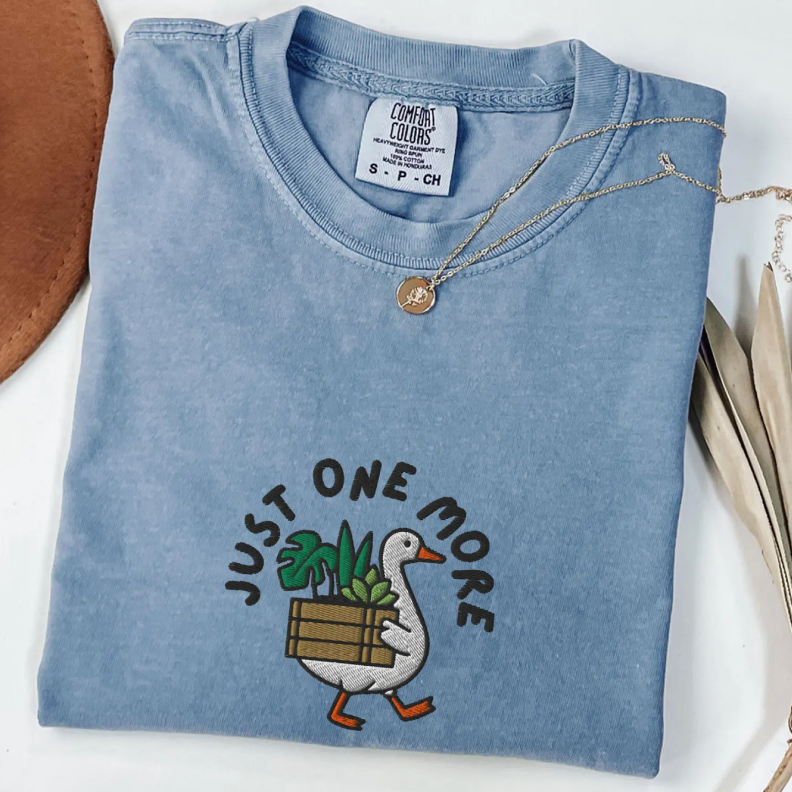 Folded blue T-shirt with a cartoon duck carrying a box of plants, surrounded by the phrase "Just One More."