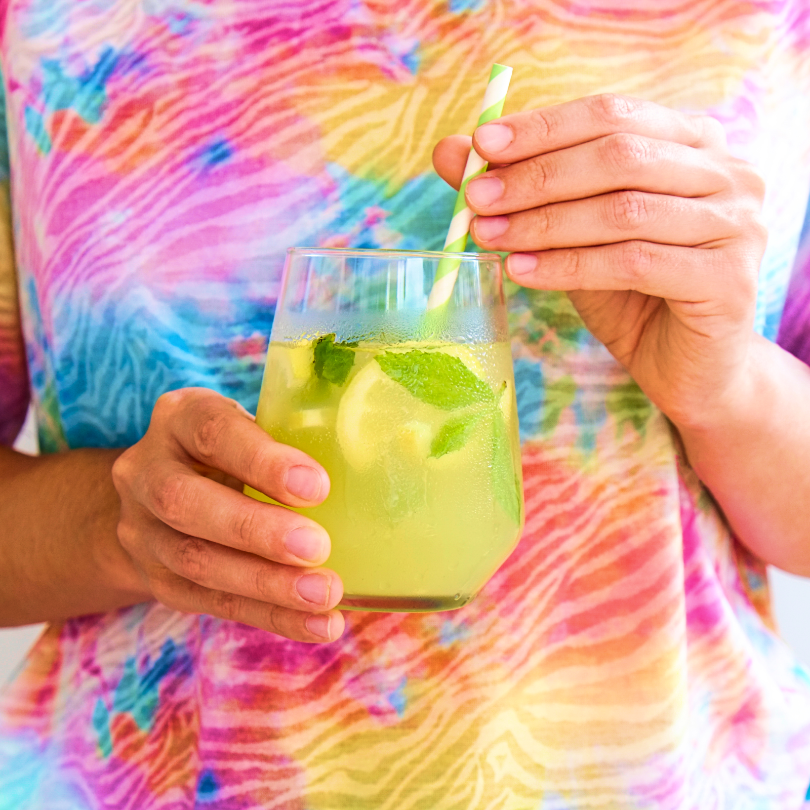 Two hands hold a glass of lime and mint-infused drink with a striped straw, against a vibrant rainbow-colored tie-dye shirt backdrop.